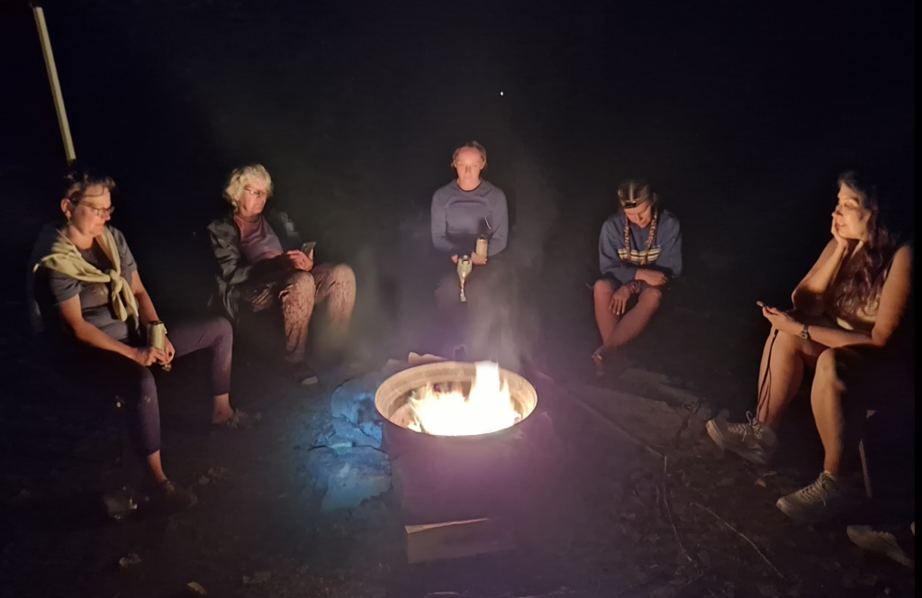 five women sit around a campfire in the dark, the glow illuminating their faces 