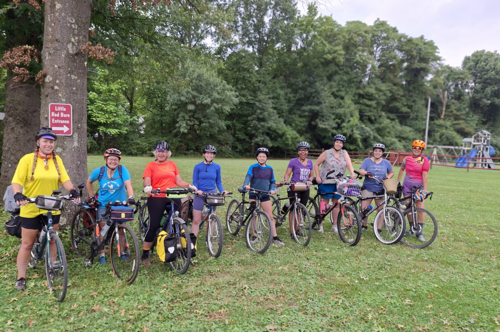 A row of women smile at the camera, standing over their bikes in a green-grass park with tall trees in the background 