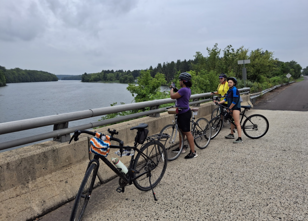 Three women stand on a bridge taking photos of the water. They have bicycles and there is an empty bike in the foreground 