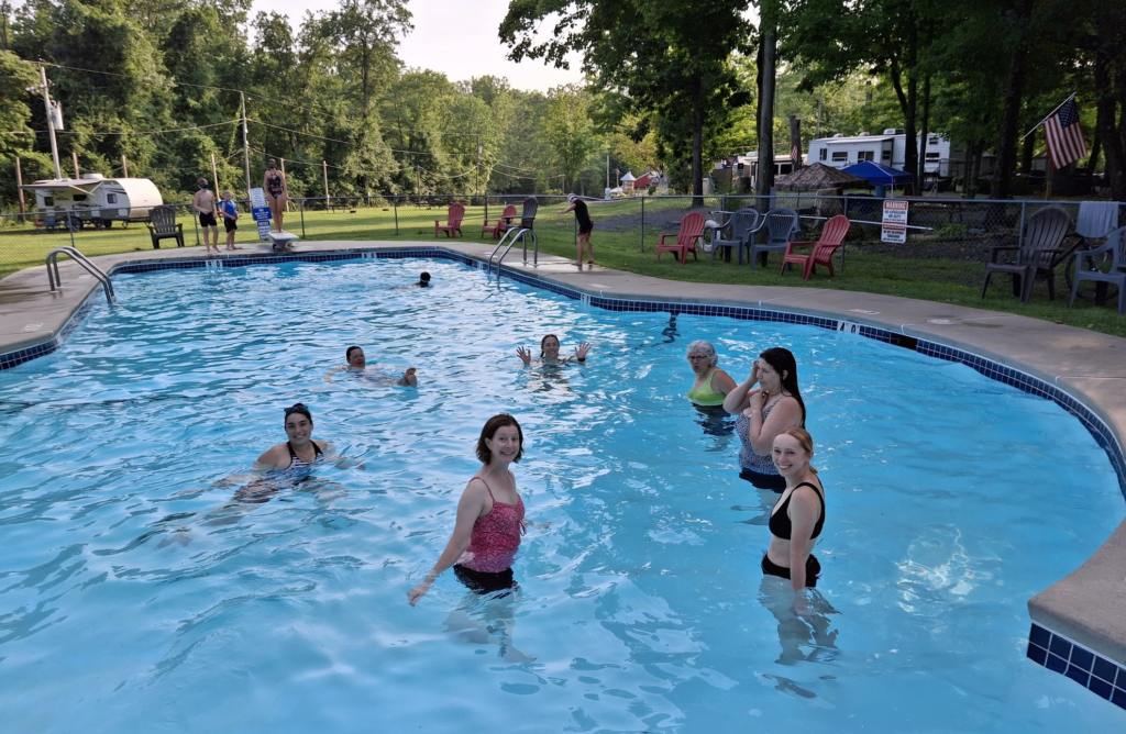 A group of people smile at the camera from a swimming pool with green grass and trees in the background 