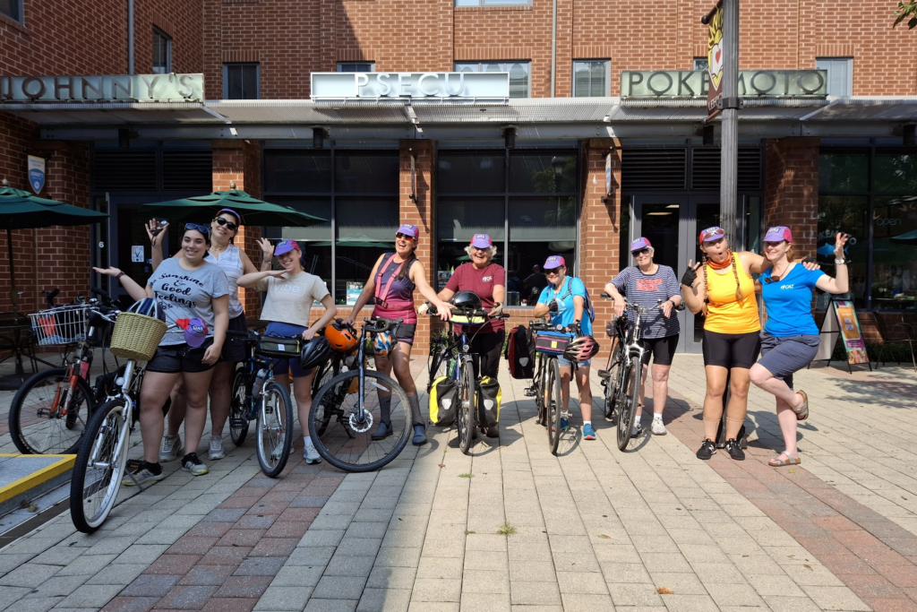 a group of women stand over bikes in front of a brick building. They are smiling and looking at the camera 