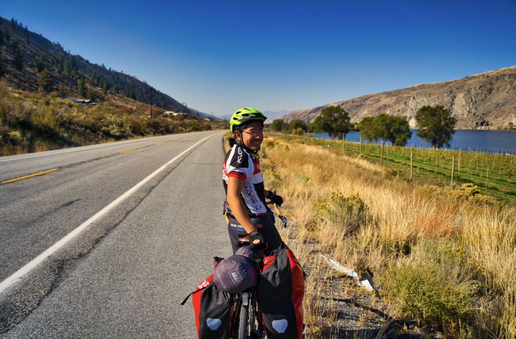 Person on a loaded bike smiling on the side of a paved road that goes along a vineyard and then river.