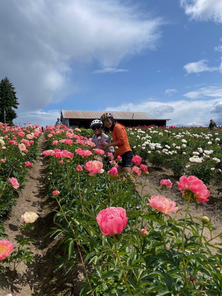 Two people in bike helmets smile with a row of pink peonies.