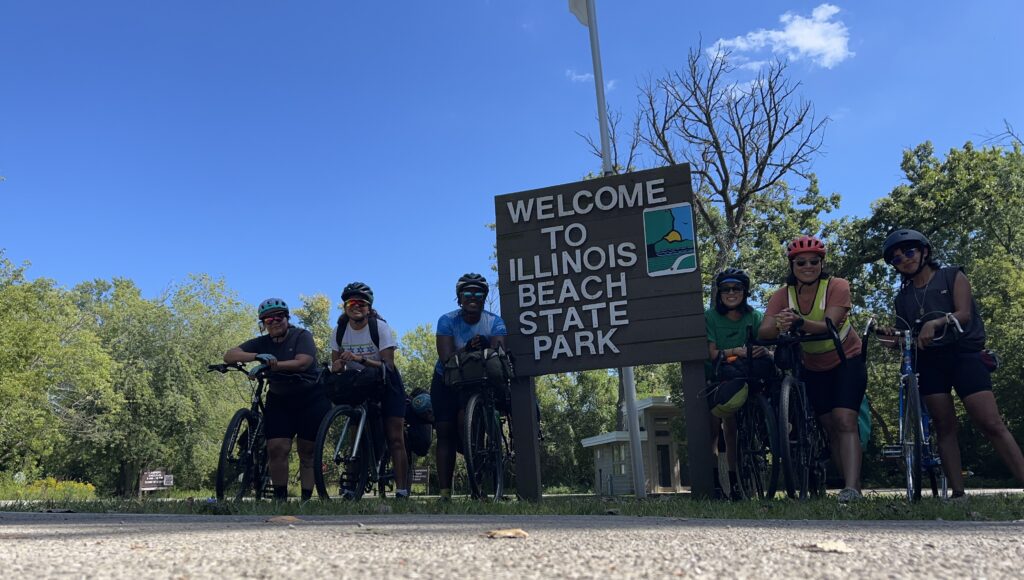 A small group of cyclists stand in front of a sign for the illinois beach state park with a blue sky and trees in the background 