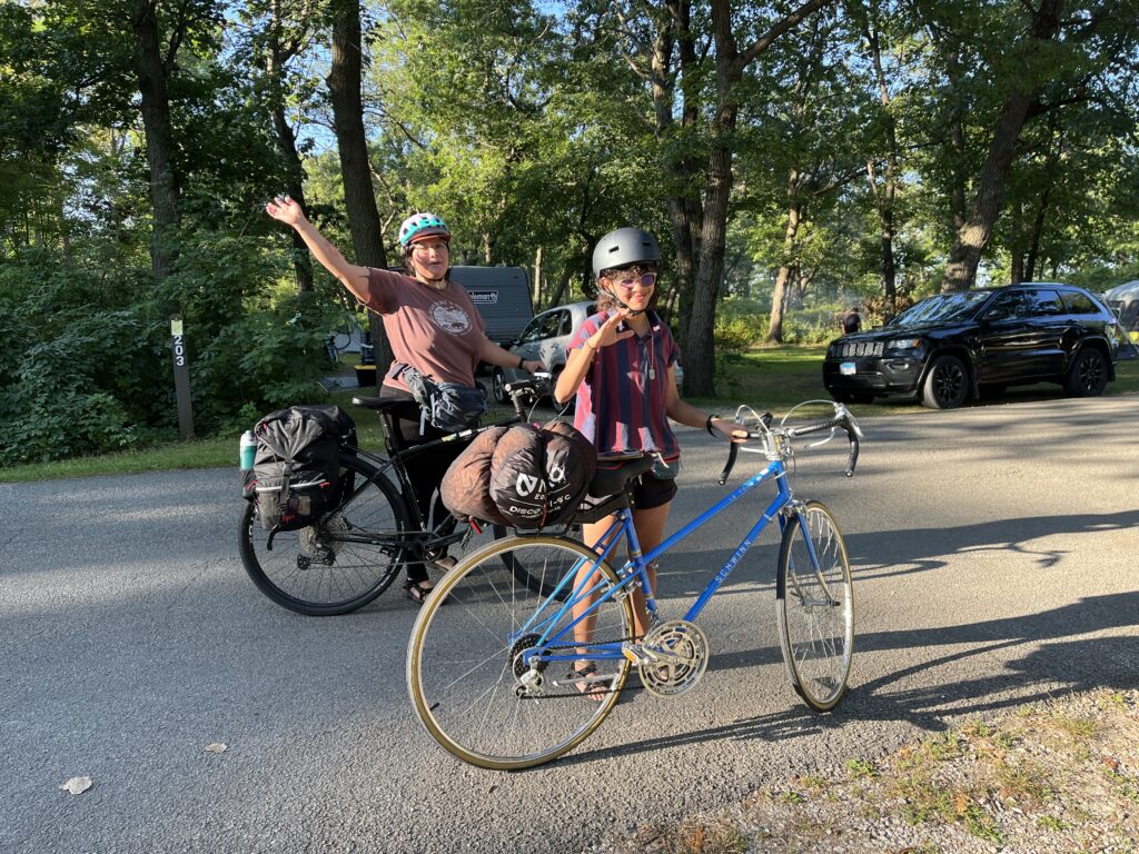 two cyclists stand with their bikes in a parking lot with trees in the background and one car. They are smiling and waving at the camera 