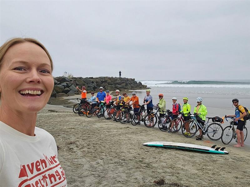 Woman smiling taking a selfie showing a large group of cyclists at the edge of the water on the beach.