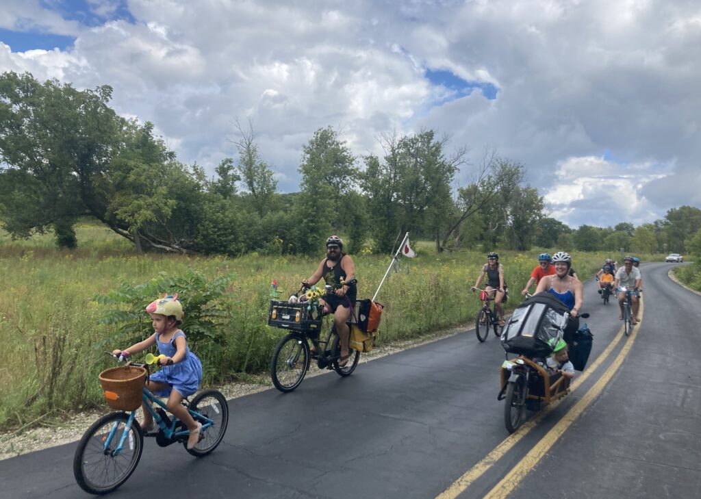 A group of cyclists young and old ride to the left of the camera on a paved road. some cyclists are on adaptive bikes