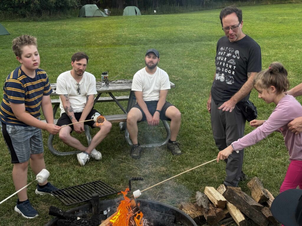 a group of people sit around a campfire in a green field roasting marshmallows 