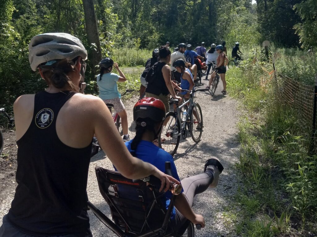 A crowd of cyclists seen from behind, some walking bikes and others on adaptive bikes on a path between brush and trees