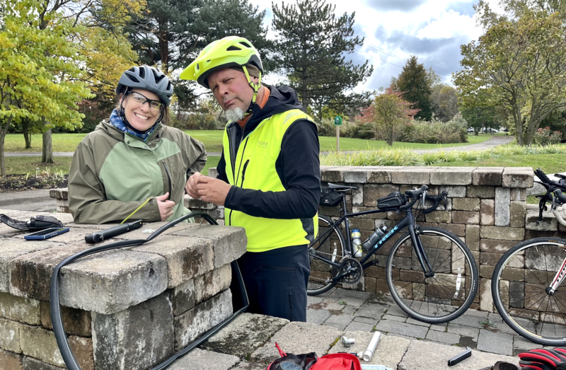 two cyclists smile at the camera, they look like they are changing a flat tire 