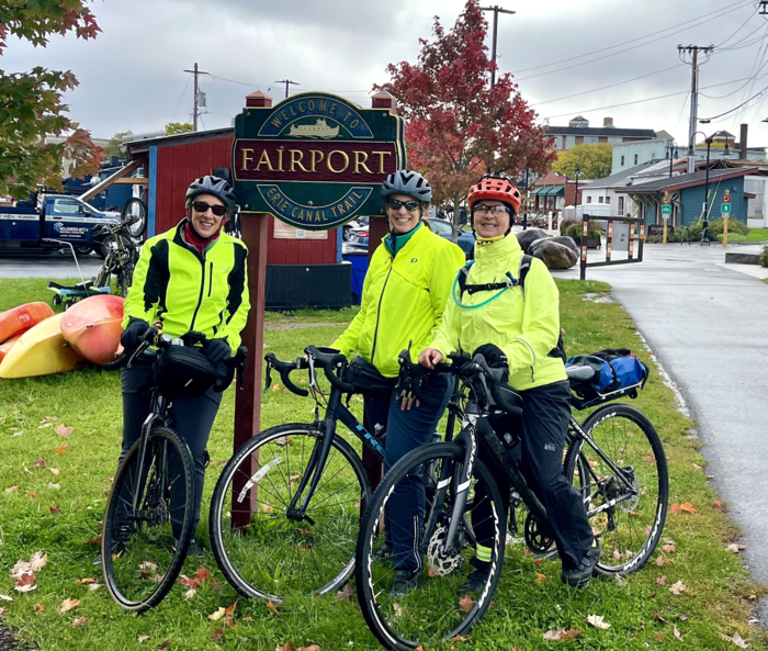 three cyclists stand in front of a sign with the words "fairmont" written on it. They are in reflective gear and the sky is gray