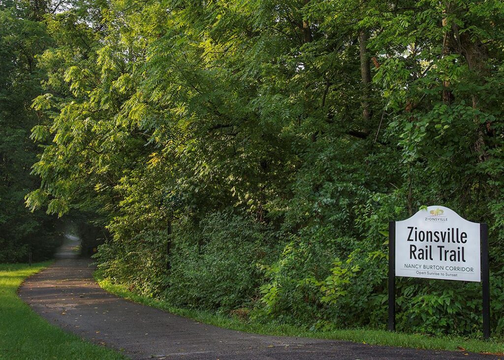 Wooded paved path with a sign that says Zionsville Rail Trail