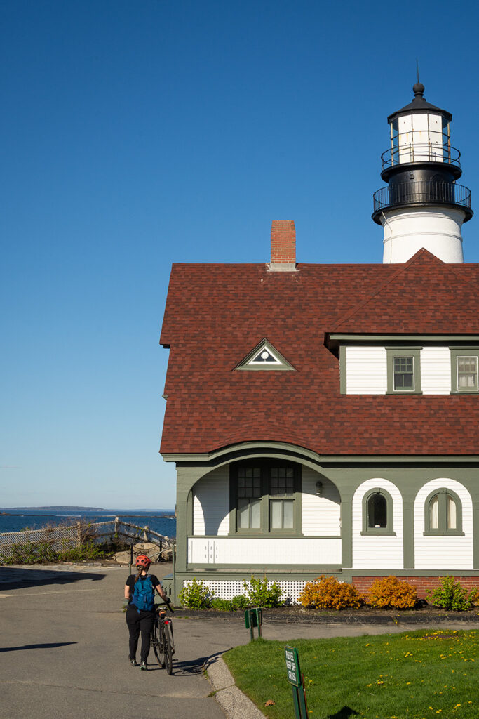 A woman walks her bike toward an old house with the top of a lighthouse peeking up behind the house.